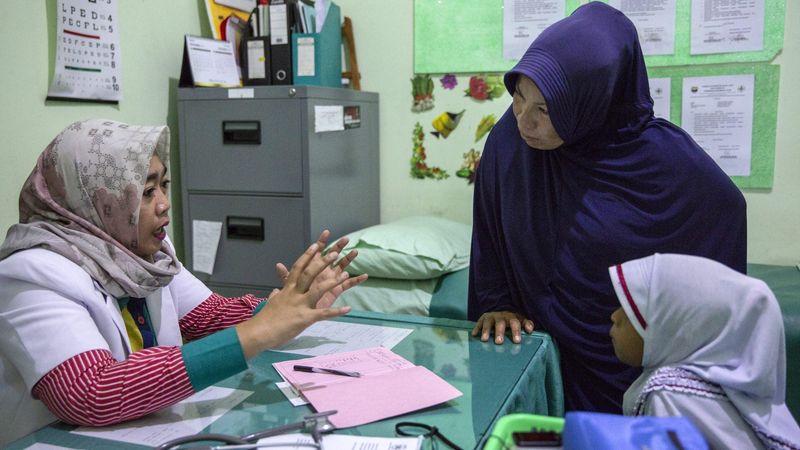 A healthcare professional consulting with a woman in a blue hijab and a young girl in a medical room, with a stethoscope on the desk