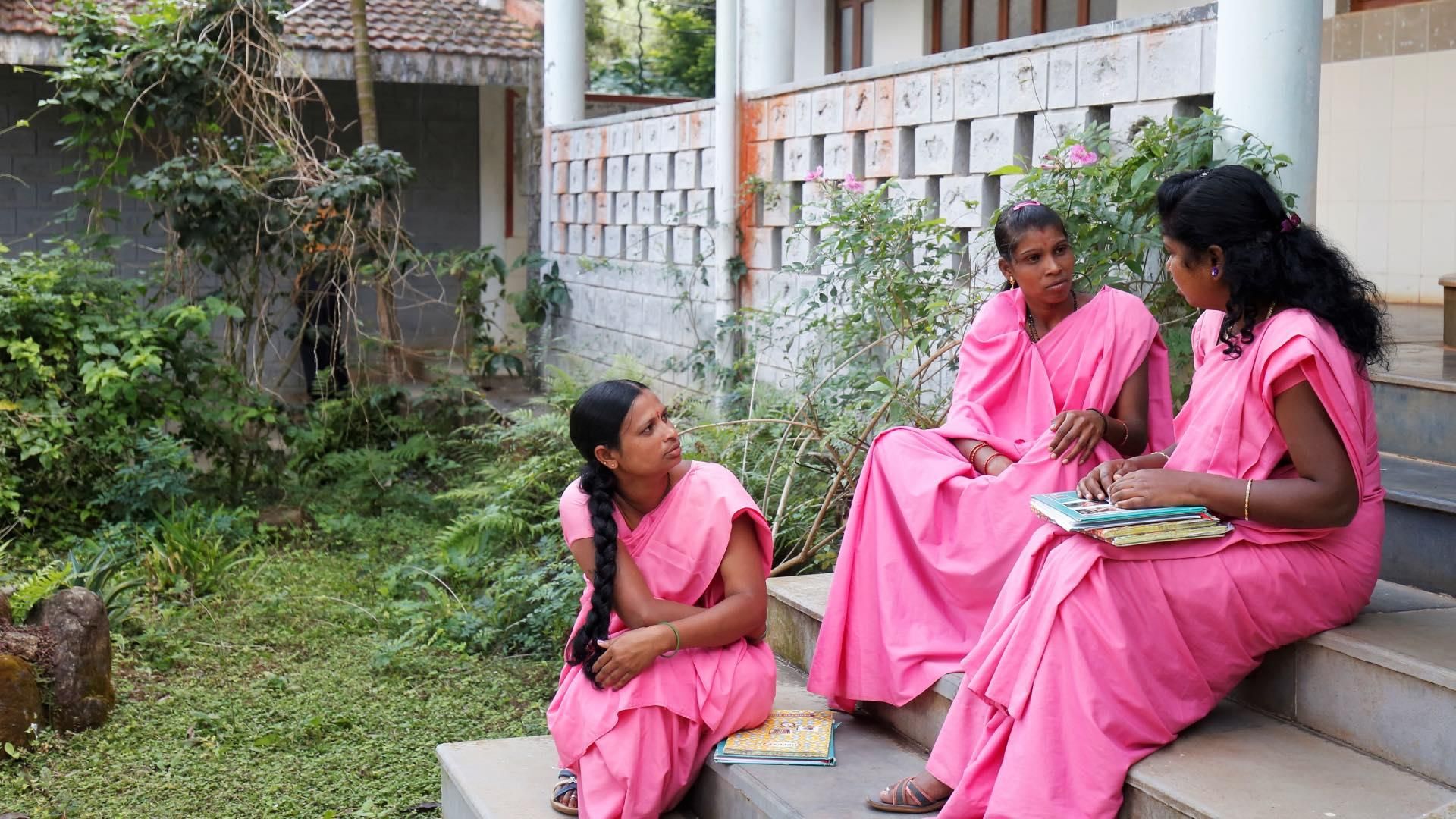 Three young women wearing traditional pink sarees sitting on stone steps in front of a rustic building, one holding books