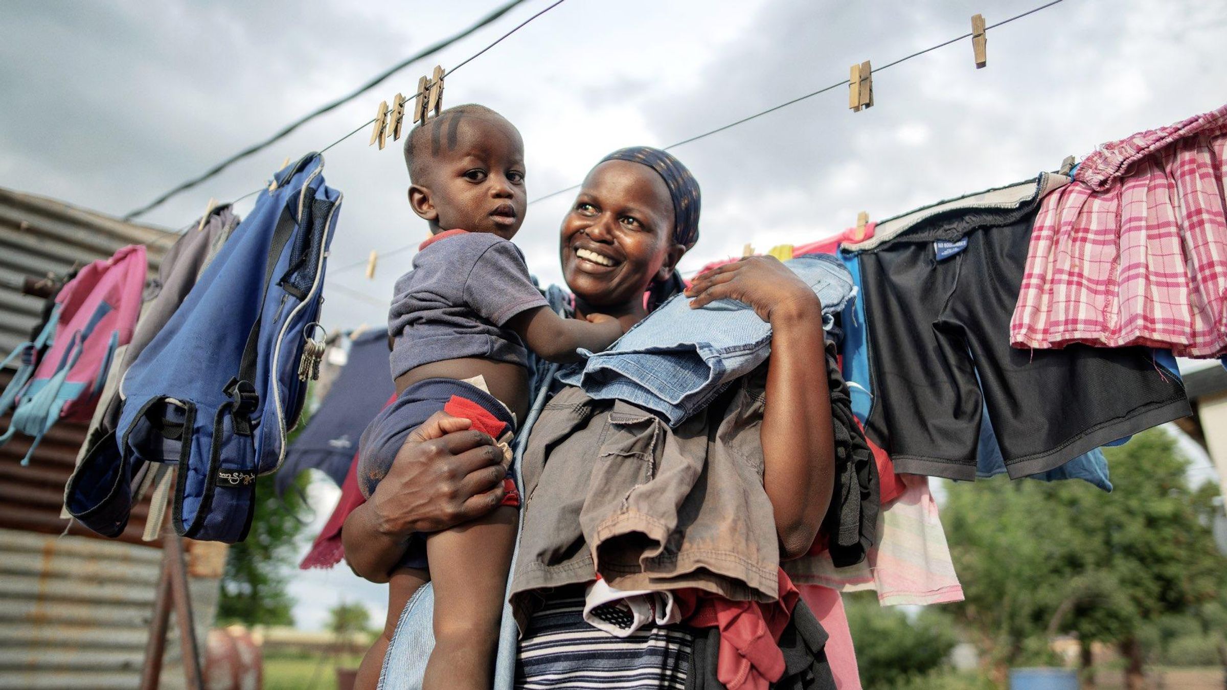A smiling woman in a striped shirt carrying a young child in her arms in front of a clothesline with drying clothes