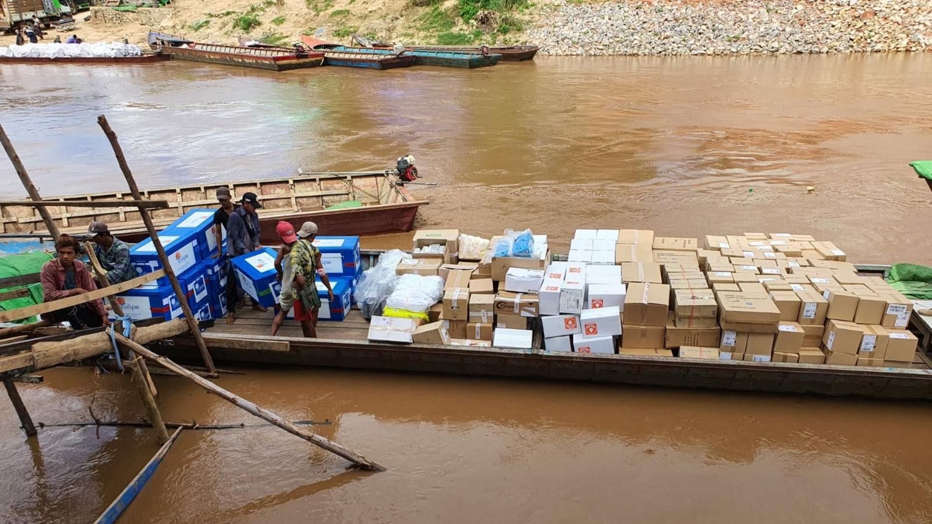 Flat-bottomed boat loaded with numerous boxes and supplies on a brown river, with people organizing humanitarian cargo