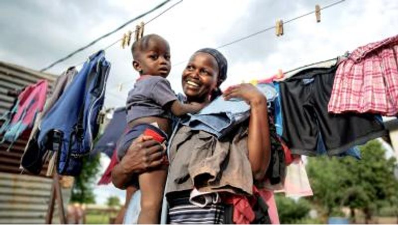 A smiling woman in a striped shirt carrying a young child in her arms in front of a clothesline with drying clothes